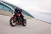 A red and black Triumph Trident motorcycle with gold accents is ridden on a curved road with a modern glass building in the background. The rider is wearing a black helmet and black riding gear.