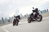 Two Triumph Trident 800 motorcycles on a curved elevated road with a city skyline in the background under a cloudy sky. The motorcycle in the foreground is black with gold accents and a rider in black gear is seated on it.