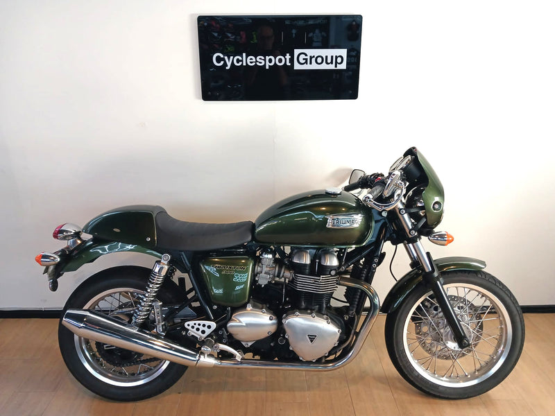 Load image into Gallery viewer, A dark green 2013 Triumph Thruxton 900 motorcycle with chrome details, parked indoors against a white wall with a 'Cyclespot Group' sign above it.
