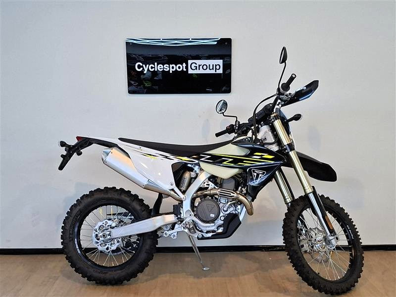Load image into Gallery viewer, A Triumph TF 450-E off-road dirt bike with a white, black, and yellow color scheme, displayed on a stand in an indoor showroom setting with a wall-mounted 'Cyclespot Group' sign behind it.

