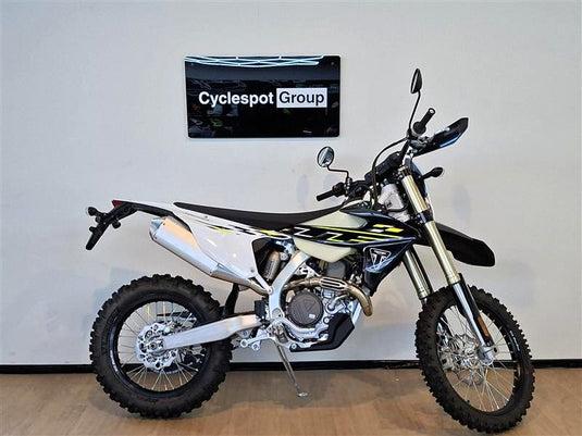 A Triumph TF 450-E off-road dirt bike with a white, black, and yellow color scheme, displayed on a stand in an indoor showroom setting with a wall-mounted 'Cyclespot Group' sign behind it.