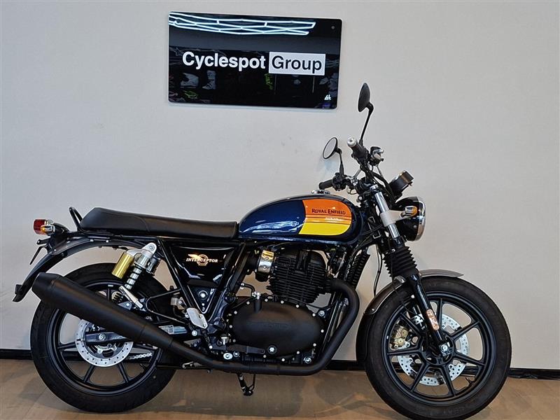 Load image into Gallery viewer, A Royal Enfield Classic Cruiser motorcycle with a black seat, black engine and exhaust, and a blue tank with orange and yellow graphics, parked indoors against a light gray wall with a 'Cyclespot Group' sign above it.
