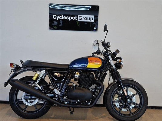 A Royal Enfield Classic Cruiser motorcycle with a black seat, black engine and exhaust, and a blue tank with orange and yellow graphics, parked indoors against a light gray wall with a 'Cyclespot Group' sign above it.