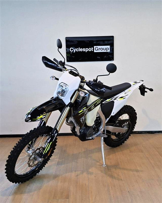 A white and black Triumph TF 250 E dirt bike with yellow accents, parked on a wooden floor against a white wall with a 'Cyclespot Group' sign.