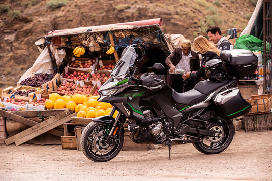 A black and green 2024 Kawasaki Versys 1000 S motorcycle with hard pannier bags is parked at a dusty outdoor market stall with people and fruit displays in the background.