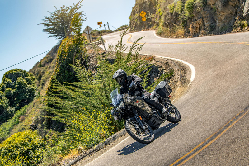 Load image into Gallery viewer, A motorcyclist wearing a black helmet and riding jacket maneuvers a 2026 Kawasaki KLR650 Adventure (LAMS) motorcycle around a sharp mountain road curve under a clear blue sky.
