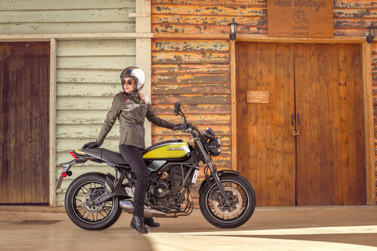 A person wearing a black leather jacket, black pants, and a white helmet sits on a yellow and black Kawasaki Z650RS motorcycle in front of a rustic wooden building.