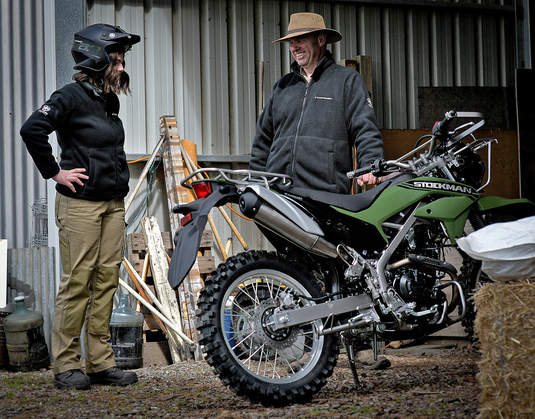 A man in a tan hat stands next to a green and black Suzuki dirt bike in a barn, while a woman in a black jacket and khaki pants wearing a motorcycle helmet looks on.