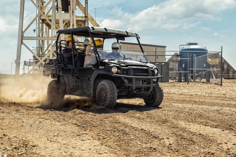 Load image into Gallery viewer, A black all-terrain utility vehicle with four passengers drives on a dusty dirt track in an industrial outdoor setting under a blue sky.
