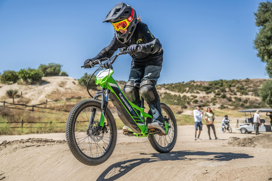 Child riding a green Kawasaki electric Elektrode bike on a dirt track with a clear blue sky and trees in the background.