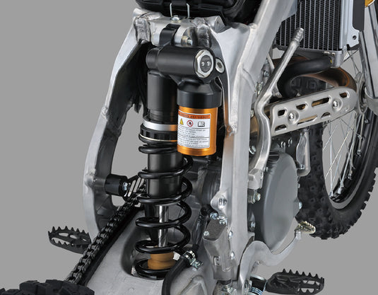 Close-up of a motorcycle's rear suspension system, featuring a black coil spring, black shock absorber, and an orange filter canister, set against a silver frame and tire.