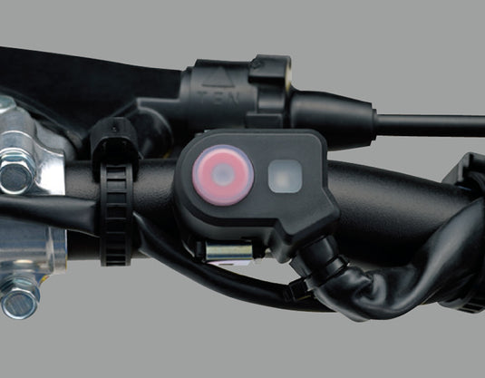A close-up of a black handlebar-mounted electronic holeshot device with a pink circular button and a small square light, attached to a motorcycle handlebar assembly against a plain gray background.