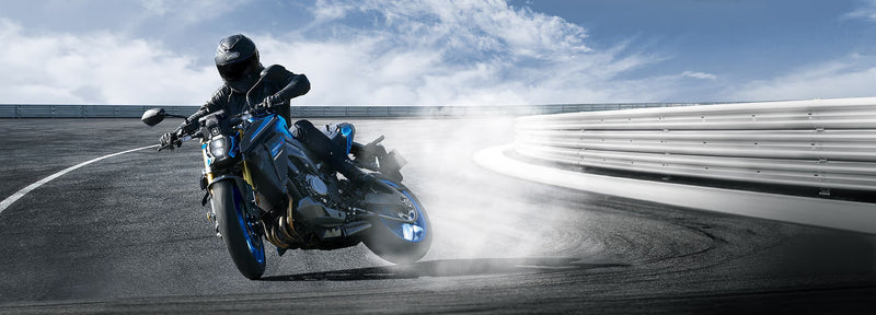 Load image into Gallery viewer, A blue and black 2026 Suzuki GSX-S 1000 RQ motorcycle with a rider in full black gear leaning into a turn on a racetrack, kicking up dust and smoke under a blue sky.
