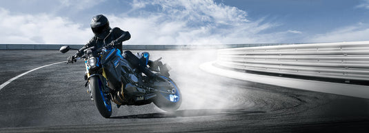 A blue and black 2026 Suzuki GSX-S 1000 RQ motorcycle with a rider in full black gear leaning into a turn on a racetrack, kicking up dust and smoke under a blue sky.
