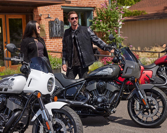 A man and a woman in black leather jackets stand outdoors beside a black Indian Sport Chief motorcycle, with other motorcycles partially visible in the foreground and a brick building in the background.