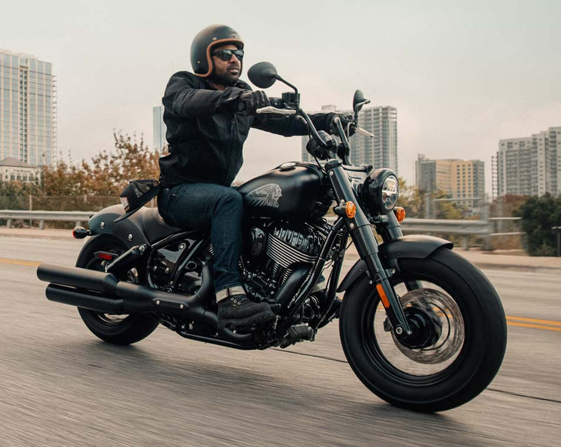 Load image into Gallery viewer, A man wearing a black helmet, black jacket, and blue jeans rides a black Indian Chief Bobber Dark Horse cruiser motorcycle on a highway with city buildings in the background.
