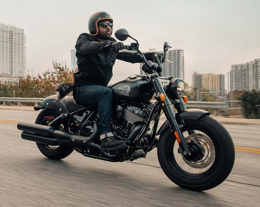 A man wearing a black helmet, black jacket, and blue jeans rides a black Indian Chief Bobber Dark Horse cruiser motorcycle on a highway with city buildings in the background.