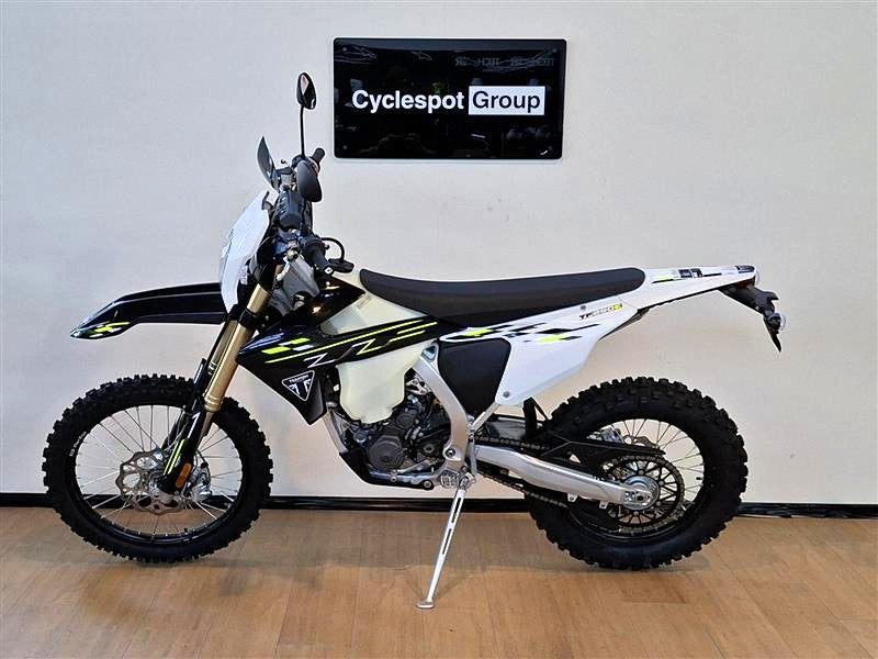 Load image into Gallery viewer, A Triumph TF 250 E dirt bike in white, black, and yellow graphics, parked on a wooden floor against a white wall with a 'Cyclespot Group' sign above it.
