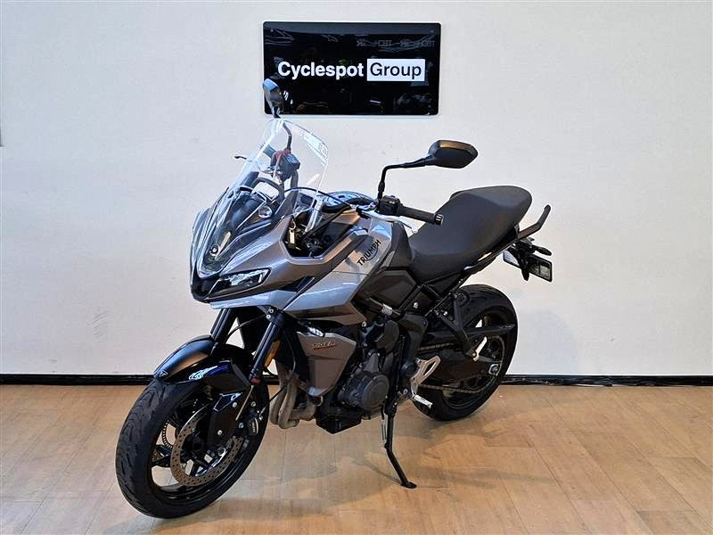Load image into Gallery viewer, A grey  2023 Triumph Tiger Sport 660 motorcycle is parked indoors on a wooden floor against a white wall, with a 'Cyclespot Group' sign visible on the wall behind it.
