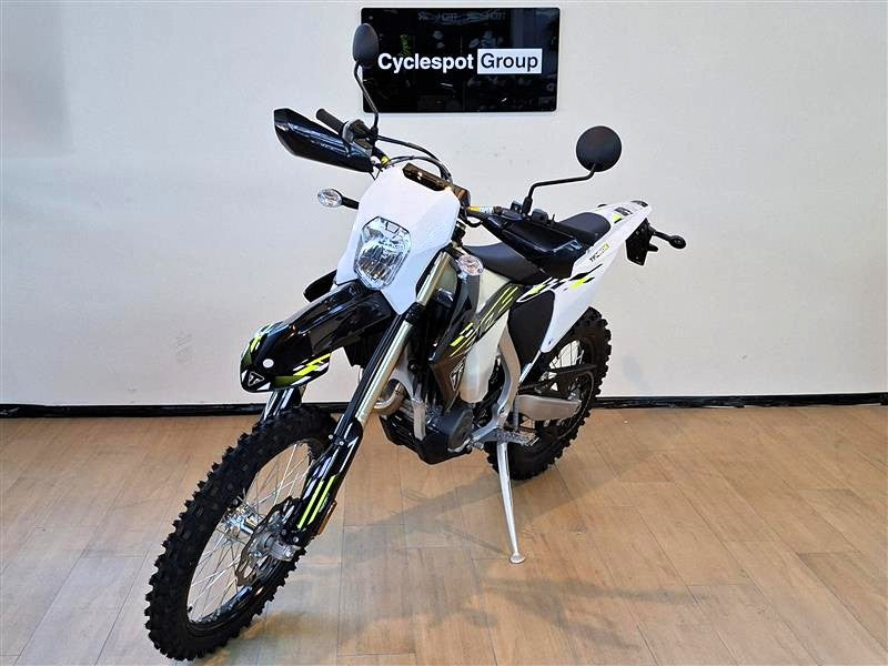Load image into Gallery viewer, A white and black Triumph TF 450-E dirt bike with yellow accents, standing on its kickstand in an indoor showroom with a wood floor and a wall-mounted sign that reads 'Cyclespot Group'.
