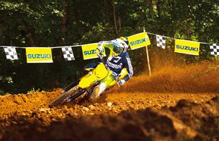 Load image into Gallery viewer, A yellow and blue 2026 Suzuki RM-Z450 dirt bike racer leans sharply into a turn on a dirt track, with a large cloud of orange-brown dust behind the bike and Suzuki banners hanging in the background.
