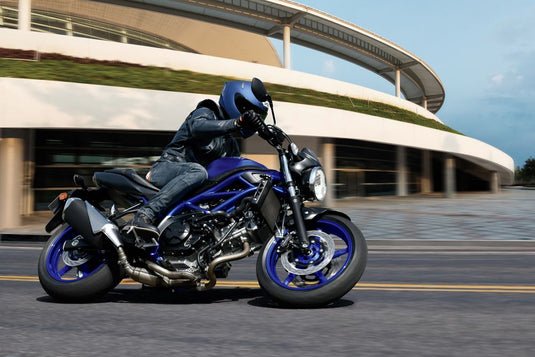 A blue and silver 2025 Suzuki SV 650 A motorcycle with a rider in a black jacket and blue helmet, captured in motion on a paved road with a modern building in the background.