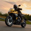 A person wearing a black helmet and riding gear sits on a yellow and black Triumph Tiger 800 motorcycle, captured from a low angle on a road during a warm sunset sky.