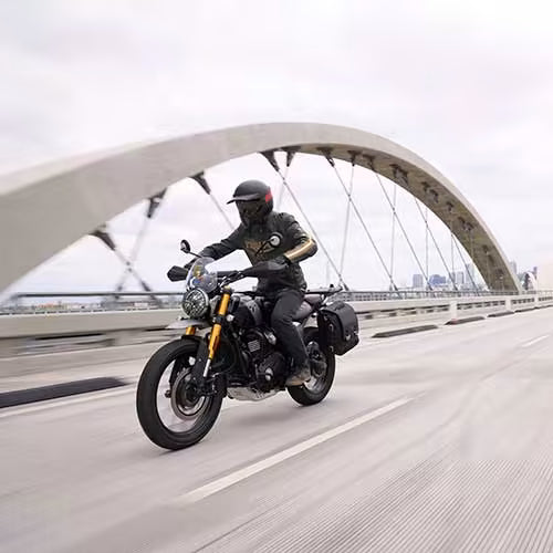 Load image into Gallery viewer, A motorcyclist wearing a black helmet and black riding gear with gold stripes rides a black and gold Triumph Scrambler 400 motorcycle on a bridge. The motorcycle has black side panniers.

