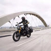 A motorcyclist wearing a black helmet and black riding gear with gold stripes rides a black and gold Triumph Scrambler 400 motorcycle on a bridge. The motorcycle has black side panniers.