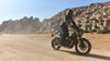 A person wearing a black riding suit and helmet rides a black and gold Triumph Scrambler motorcycle motorcycle on a dusty desert road with large rock formations in the background.