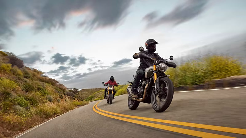 Load image into Gallery viewer, A motorcycle rider in black gear rides a black  Triumph Scrambler 400 motorcycle on a winding road, with another rider and motorcycle visible in the background under a dramatic cloudy sky.
